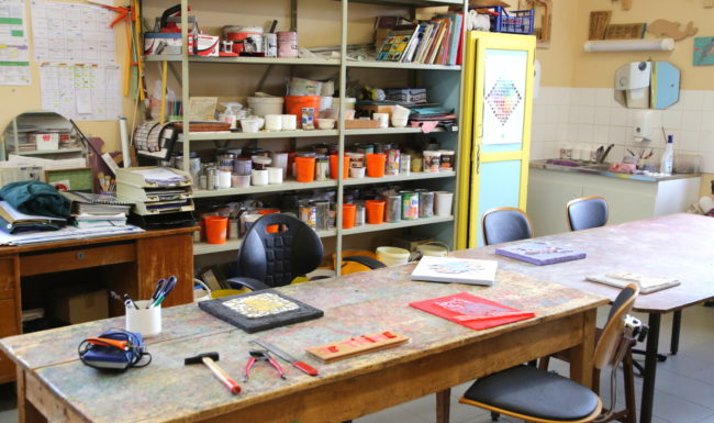 photo de l'Atelier mosaïque de la providence, étagères remplies de matériels et table de travail avec des travaux réalisés par des jeunes de l'établissement